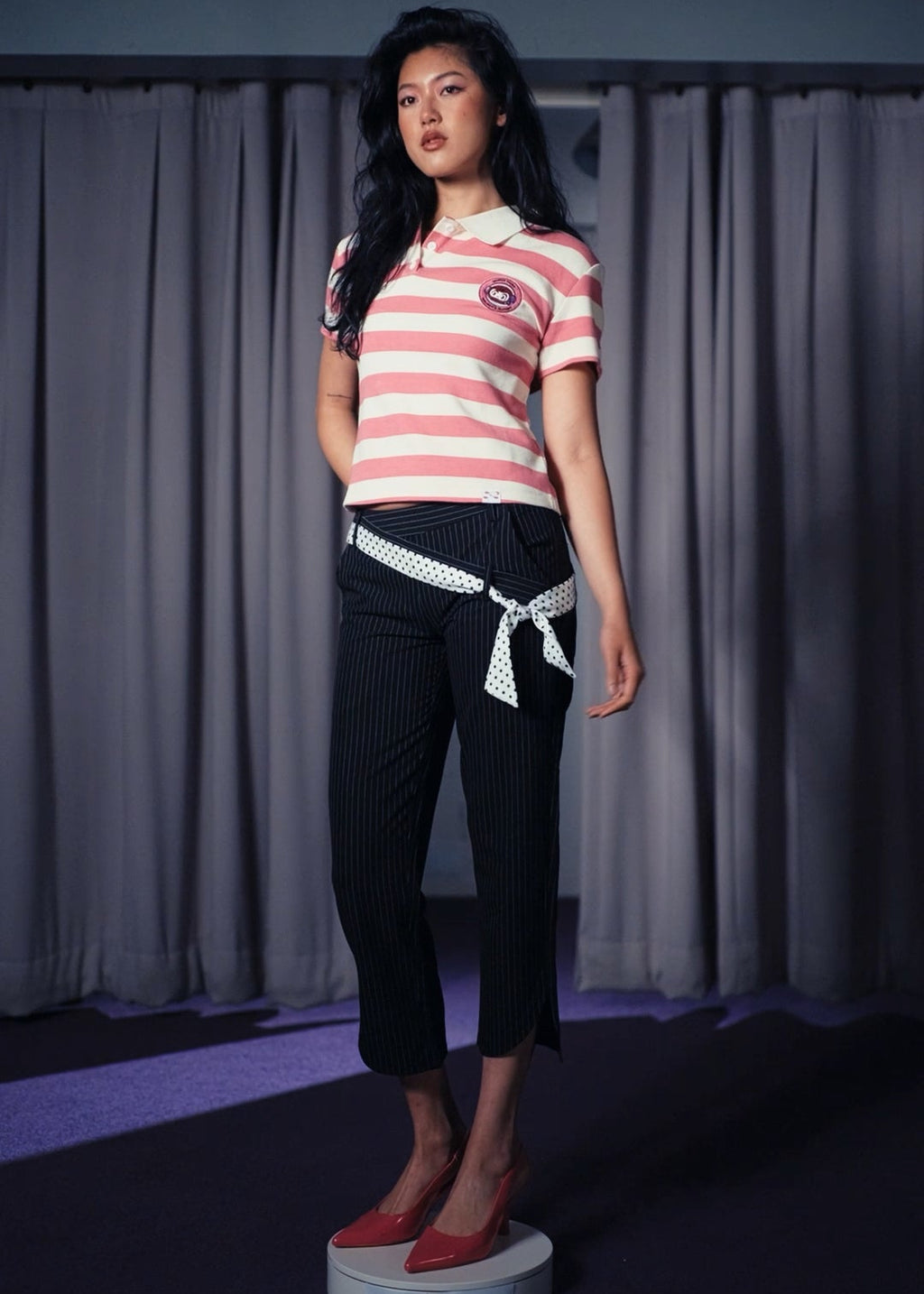 Woman wearing a striped shirt and black pants with a white belt, standing against a gray curtain.