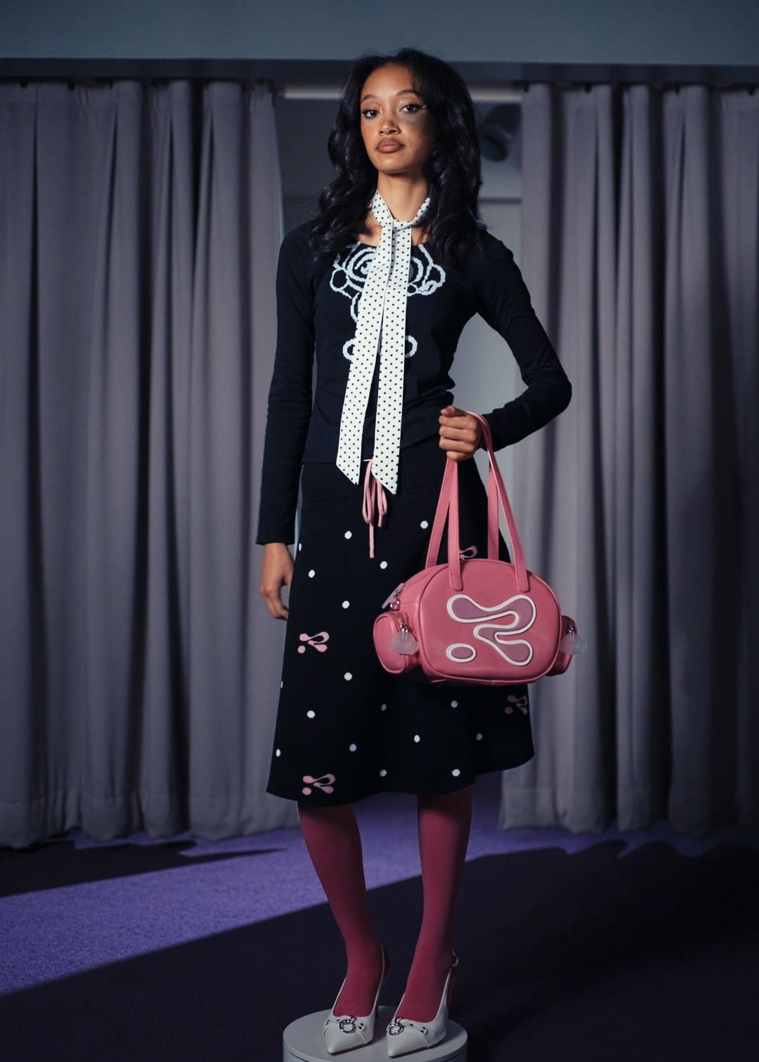 Woman holding a pink handbag with a brand logo against a curtain background